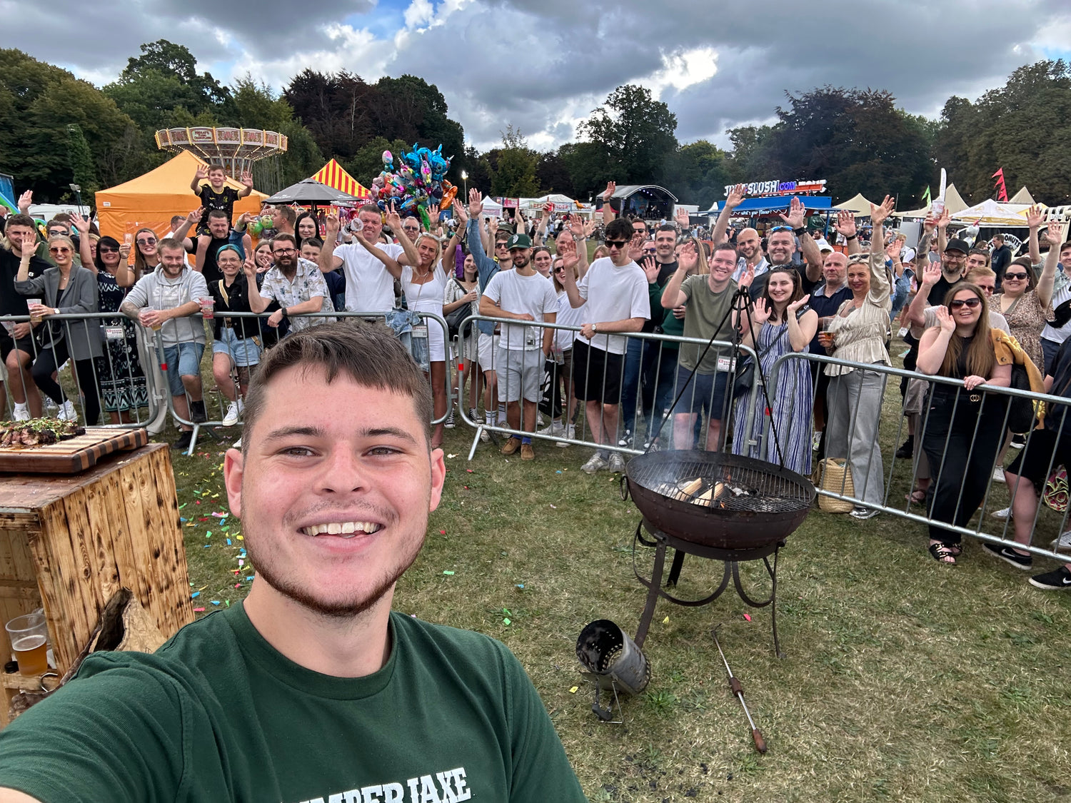 A young man in a green 'Lumberjaxe' t-shirt takes a selfie in front of a large, excited crowd at an outdoor festival. The crowd, diverse in age and attire, stands behind a metal barrier, waving and smiling. In the foreground, a rustic wooden table holds food, and a round charcoal grill with flames burning is set up on the grass. The festival setting features colorful tents, fairground rides, food stalls, and balloons in the background under a cloudy sky.