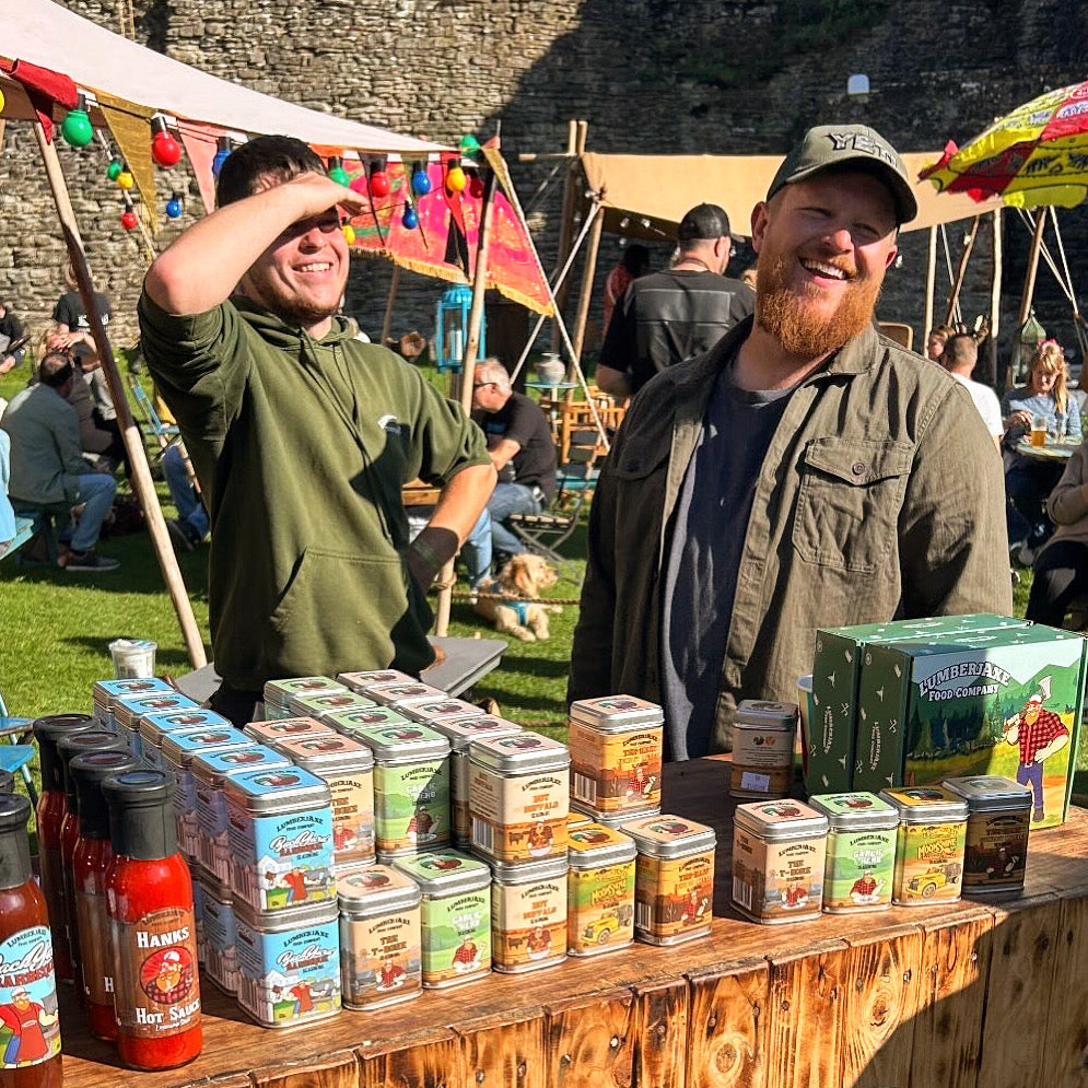 Two men stand behind a rustic wooden market stall displaying an array of 'Lumberjaxe' seasoning tins and hot sauce bottles at an outdoor festival. One man, wearing a green hoodie, shields his eyes from the sun while smiling, while the other, a bearded man in a dark green jacket and YETI cap, laughs. The setting features colorful tents, string lights, umbrellas, and people enjoying the event against the backdrop of an old stone wall. A dog rests on the grass in the background.