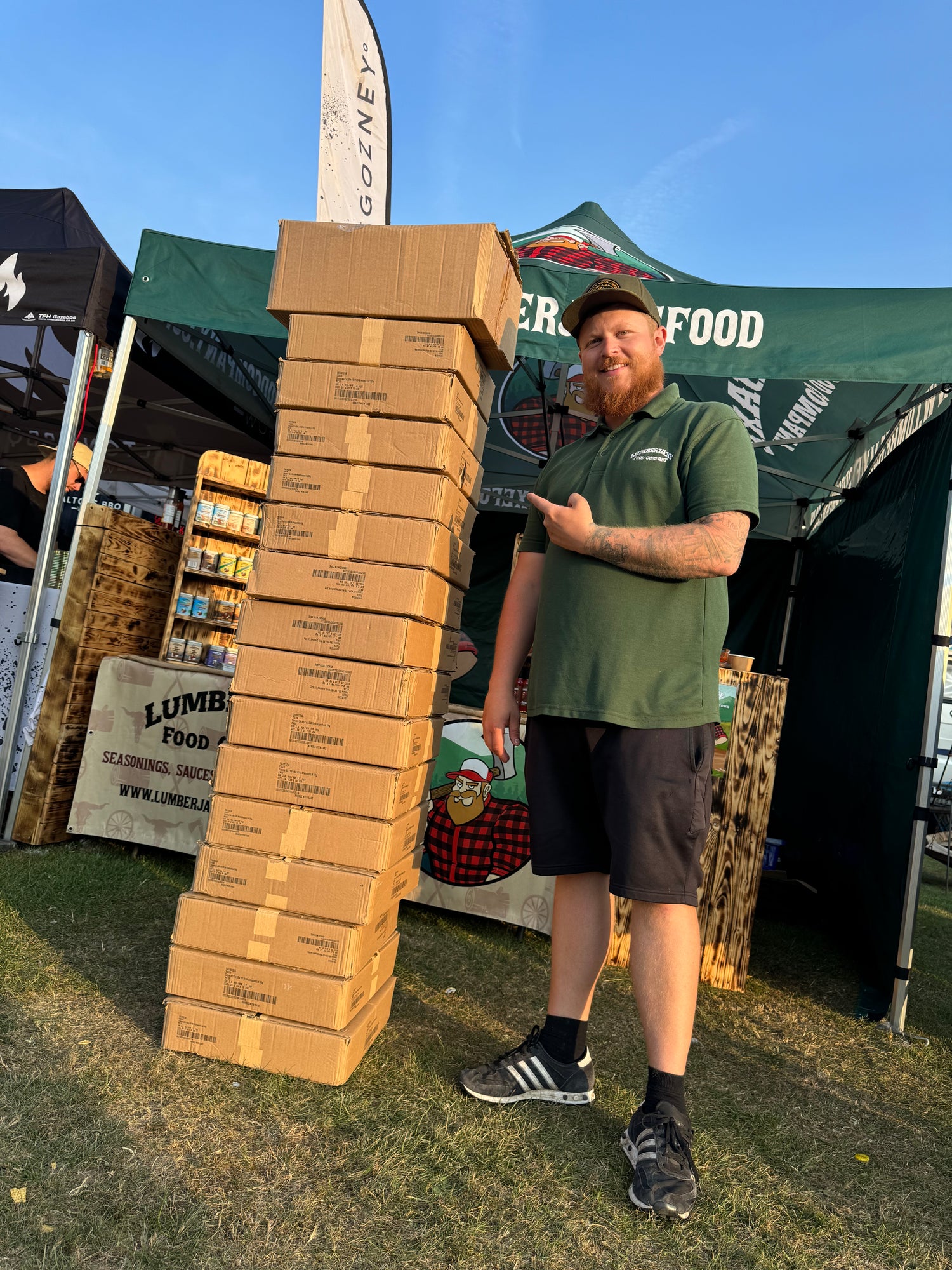 A bearded man wearing a green 'Lumberjaxe' t-shirt and black shorts stands outdoors at a festival, pointing at a tall, slightly leaning stack of cardboard boxes. He has a confident smile and is wearing a black cap and Adidas sneakers. Behind him, a green 'Lumberjaxe' tent is set up, displaying seasonings, sauces, and food products. The setup includes wooden display crates, banners with a lumberjack character, and other vendor tents visible under a clear blue sky.
